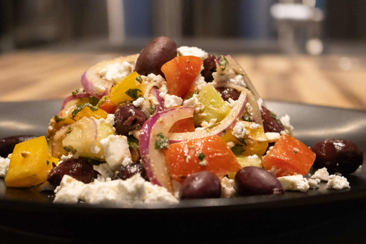 A mound of Greek Salad with diced peppers, red onion, kalamata olives, and crumbled feta on a dark plate.
