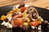 Greek Salad on a black plate with peppers, red onion, black olives, and crumbled feta cheese.