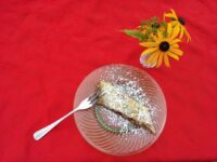 A slice of Apple Strudel dusted with powdered sugar on a glass plate with a fork, beside yellow flowers.