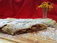 Sliced Apple Strudel on a wooden board, dusted with powdered sugar, fruit filling visible in cross-section.