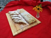 Sliced Apple Strudel dusted with powdered sugar on a wooden board, red cloth and yellow flowers behind