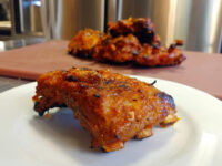 A charred, glazed Czech BBQ – Honey Pork Ribs piece on a white plate, more ribs on a board behind.
