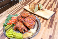 Glazed Czech BBQ – Honey Pork Ribs on a metal plate with pickles, tomatoes, greens, and mustard.