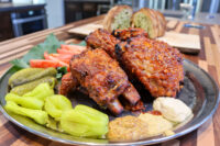 Glazed Czech BBQ – Honey Pork Ribs on a metal plate with pickles, peppers, tomato, and two dips.