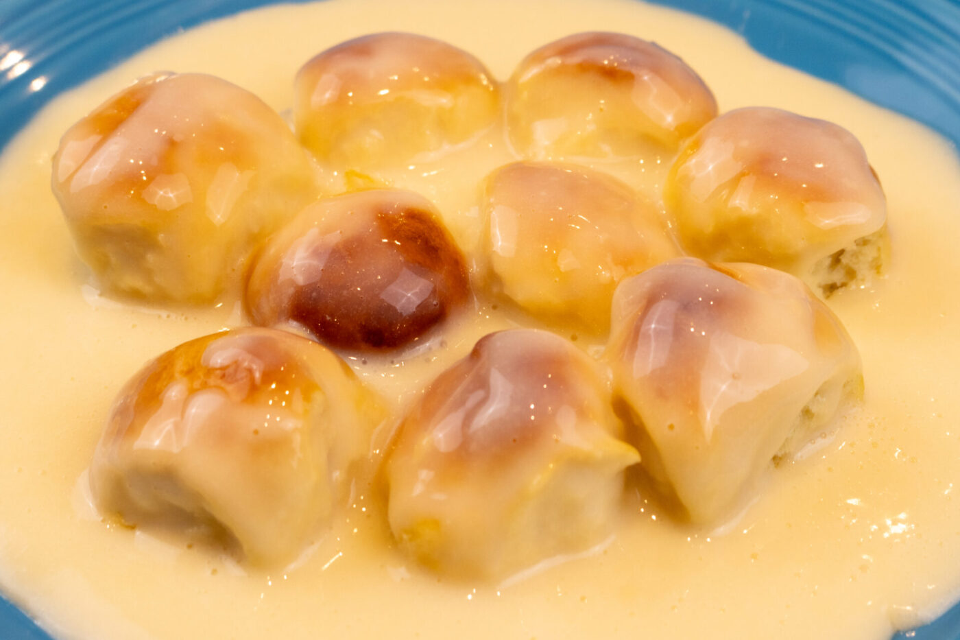 Nine golden-browned Little Buns with Vanilla Pudding (Buchticky s Krémem) sitting in custard sauce on a blue plate