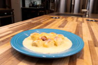 Small round buns of Little Buns with Vanilla Pudding (Buchticky s Krémem) clustered in pale cream sauce on a blue plate atop a wooden counter.