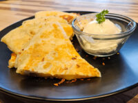 Golden Quesadilla wedges on a black plate next to a glass bowl of sour cream garnished with parsley.