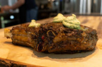 Cowboy Steak Seared bone-in Cowboy Steak topped with three dollops of green herb butter on a wooden board.