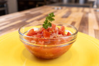 Glass bowl of Czech Tomato Salad with Onion, garnished with a sprig of herb, on a yellow plate.