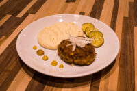 Karbenátky (Czech Burger) patty on a plate with mashed potatoes, pickle slices, and mustard dots.