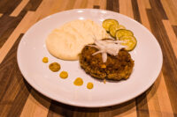 Breaded Karbenátky (Czech Burger) patty topped with onion, served with mashed potato and cucumber slices.