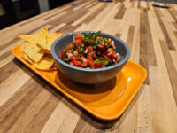 Gray bowl of chunky tomato salsa with cilantro and onion, tortilla chips on orange tray beside it.