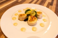 Four seared scallops with lemon twist, arugula, and dots of spicy sauce on a white plate.