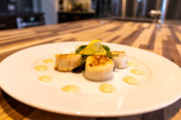 Three seared scallops with lemon and greens on a white plate, dotted with pale sauce, for Scallops with Spicy Sauce.