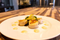 Four seared scallops with a lemon slice, arugula, and dots of spicy sauce on a white plate.