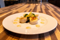 Seared scallops with spicy sauce dots, arugula, and lemon garnish on a white plate on wood table.
