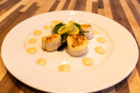 Four seared scallops with lemon, arugula, and dots of spicy sauce on a white plate
