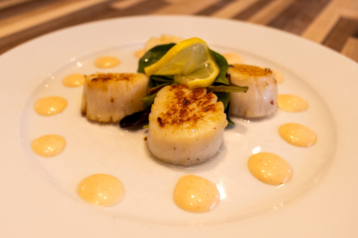 Three seared scallops with lemon twist and arugula, dots of spicy sauce ringing the plate