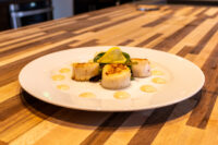 Three seared scallops with pale sauce dots, lemon and arugula garnish, Scallops with Spicy Sauce on a white plate.