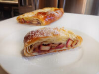Sliced Apple Strudel on a white plate showing fruit-filled layers, dusted with powdered sugar, with a second strudel behind.