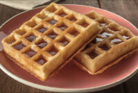 Two square Waffles drizzled with syrup, stacked on a pink plate.