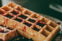 Close-up of golden Waffles with deep grid pockets, drizzled with red fruit sauce.