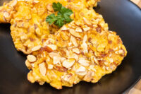 Almond Chicken Schnitzel cutlet coated in sliced almonds on a black plate, garnished with parsley.