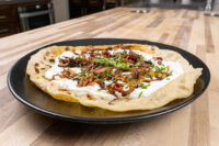 Lokše Flat Bread with Bryndza Cheese Substitute and Bacon topped with fried onions and chives on a black plate.