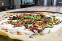 Lokše Flat Bread with Bryndza Cheese Substitute and Bacon topped with onions, bacon bits, and chives on a dark board.