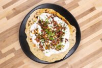 Lokše Flat Bread with Bryndza Cheese Substitute and Bacon topped with white sauce, crispy onions, bacon bits, and chives.