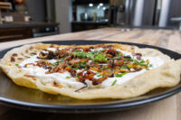 Lokše Flat Bread with Bryndza Cheese Substitute and Bacon on a black plate, topped with cream, onions, and herbs.