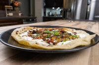 Lokše Flat Bread with Bryndza Cheese Substitute and Bacon topped with cream, crispy bits, and chives on a black plate.