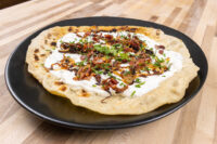 Lokše Flat Bread with Bryndza Cheese Substitute and Bacon on a black plate, topped with crispy bacon and chives.