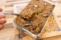 Spoon lifting a portion of Mushroom Kuba (Houbovy Kuba) with barley and mushrooms from a glass baking dish.