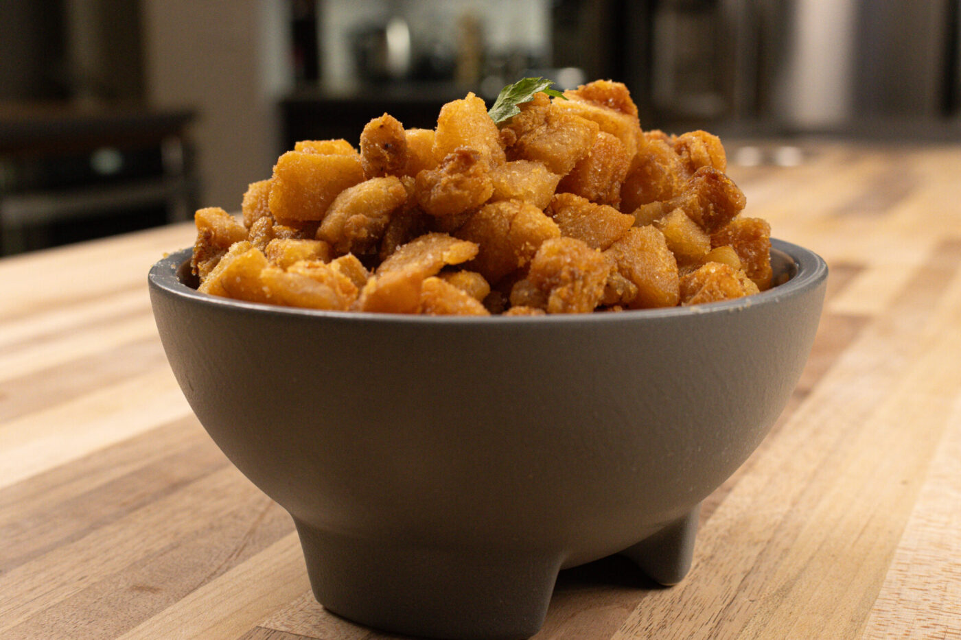 Bowl of golden-brown Pork Cracklings (Škvarky) with a green garnish leaf on a wooden table.