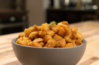 Grey bowl of golden crispy Pork Cracklings (Škvarky) topped with a herb leaf on a wooden table.