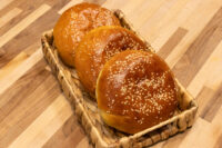 Burger Buns Three sesame-topped Burger Buns with a glossy golden crust, arranged in a woven basket on a wooden table.