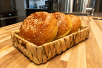 Burger Buns Three golden Burger Buns topped with sesame seeds sitting in a woven basket on a wood counter