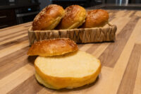 Burger Buns A sliced Burger Bun in front of a basket holding three more sesame-topped Burger Buns on a wood counter.