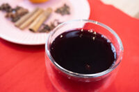 Glass of dark red Czech Mulled Wine Svarak on a red cloth, with cinnamon sticks and cloves on a plate behind it.