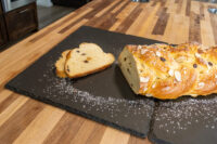 Braided Christmas Vánočka (Houska) loaf topped with sliced almonds, two cut slices showing raisins, on a slate board.