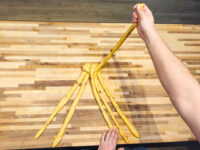 Hand pulls one dough strand upward from a knotted center as six others splay out for Christmas Vánočka shaping.