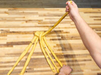 Hand braids several dough strands into a knot, shaping Christmas Vánočka (Houska) on a wooden table.