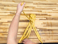 Hands braiding strands of yellow dough for Christmas Vánočka (Houska) on a wooden countertop.