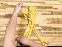 Hands braiding strands of dough with dark specks while shaping Christmas Vánočka (Houska) on a wooden board.