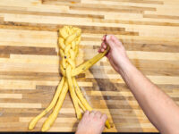 Hands braiding strands of yellow dough into Christmas Vánočka (Houska) on a wooden counter.