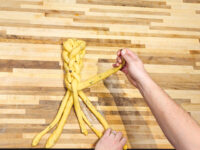Hands braiding strands of yellow dough for Christmas Vánočka (Houska) on a wooden board.
