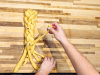 Hands shaping braided Christmas Vánočka (Houska) dough, with loose strands still unbraided on a wooden board.