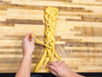 Hands braiding strands of yellow dough into a Christmas Vánočka (Houska) on a wooden board.