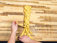 Hands braiding strands of yellow dough for Christmas Vánočka (Houska) on a wooden countertop.